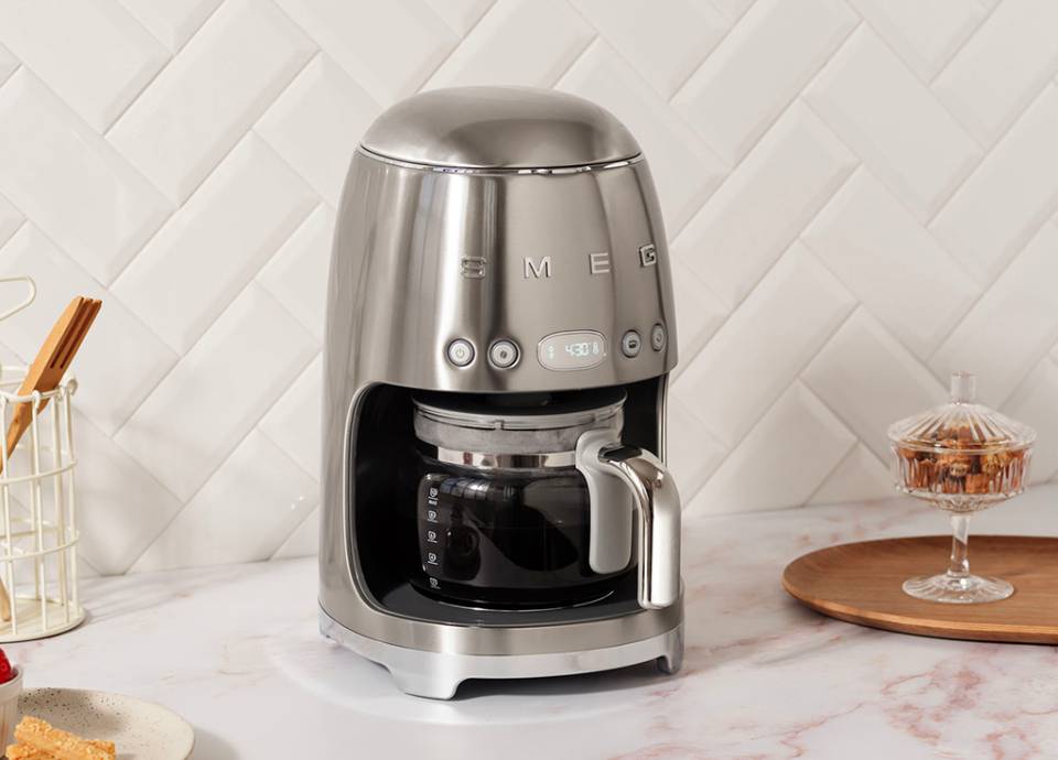 Stainless steel drip filter coffee machine with glass carafe on a light marble kitchen countertop