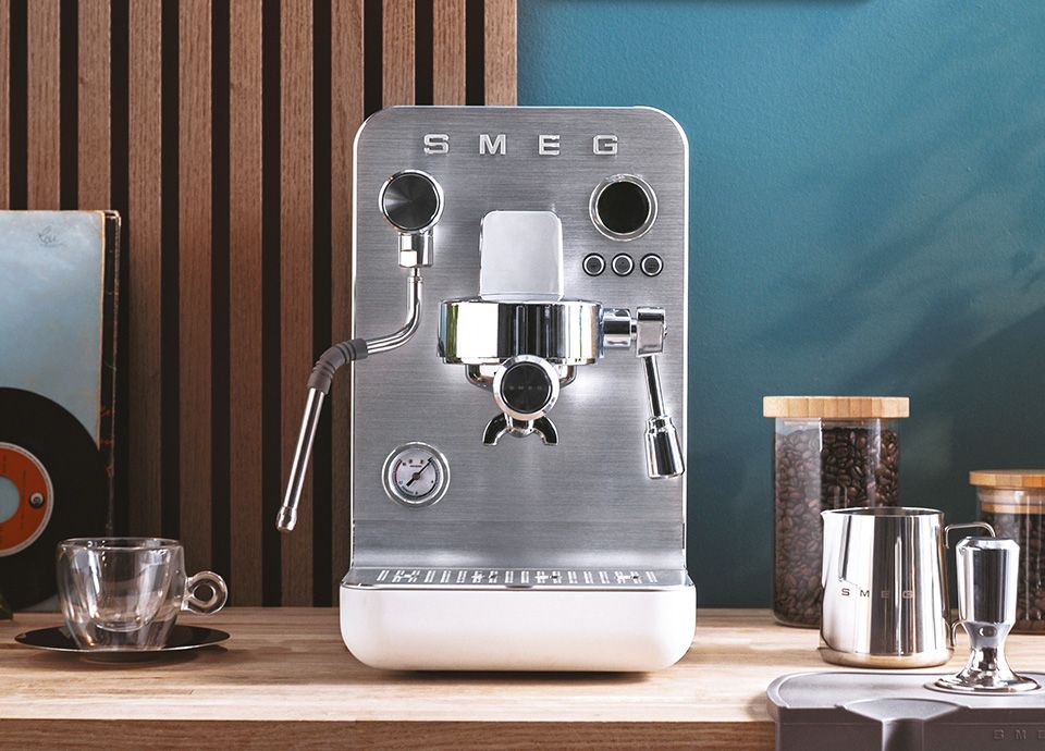 Front view of a stainless steel SMEG espresso machine on a wooden countertop, featuring a steam wand, portafilter, and pressure gauge, with a glass cup, coffee beans container, milk jug, and tamper arranged beside it against a modern kitchen backdrop.
