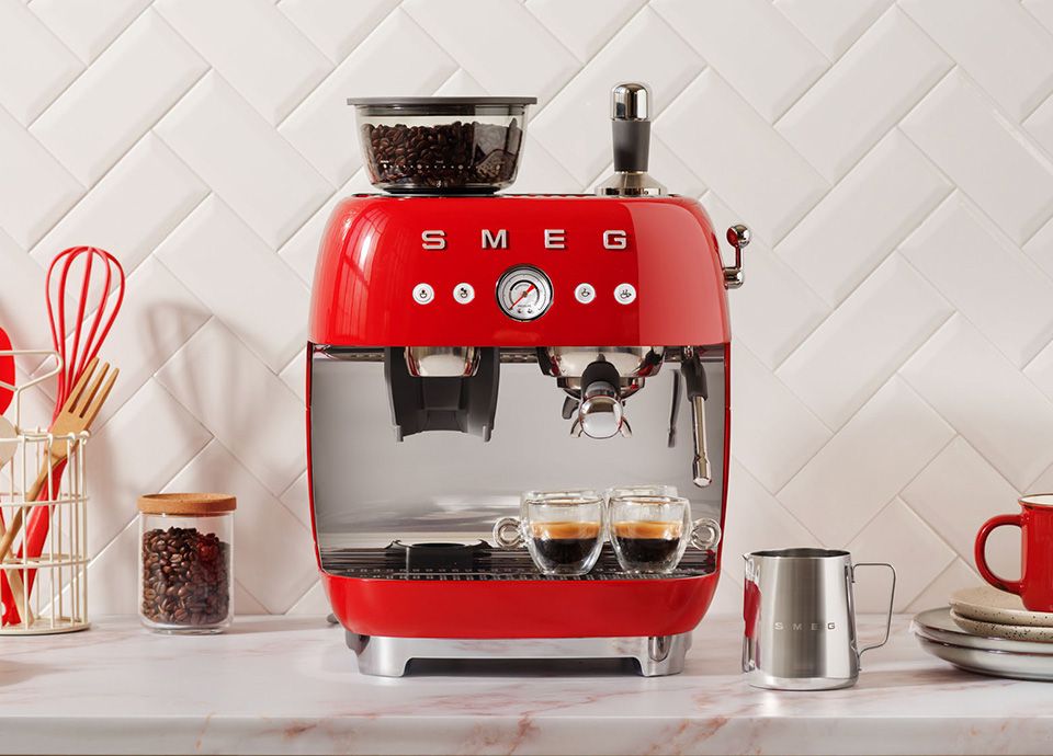 Red Smeg espresso machine with built-in grinder, dual glass cups brewing coffee, and stainless steel milk jug on a kitchen countertop