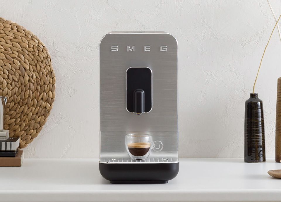 Brushed stainless steel Smeg espresso machine with a glass cup of espresso on a minimalist countertop