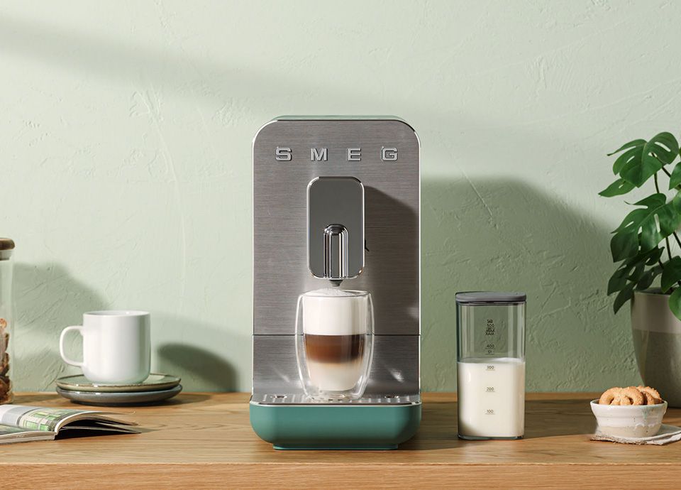 Smeg coffee machine preparing a layered latte with milk foam, placed on a wooden countertop with a milk container and kitchen accessories