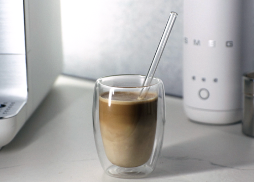 Glass of classic caffè latte with layered espresso and warm milk, served in a double‑wall glass with a glass straw.