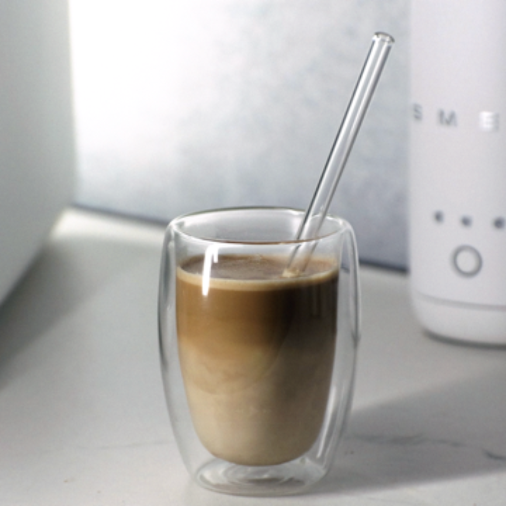 Glass of classic caffè latte with layered espresso and warm milk, served in a double‑wall glass with a glass straw.