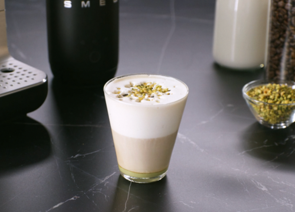 Glass of pistachio latte topped with milk foam and chopped pistachios on a dark countertop.
