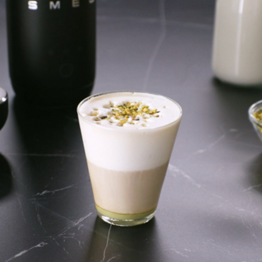Glass of pistachio latte topped with milk foam and chopped pistachios on a dark countertop.