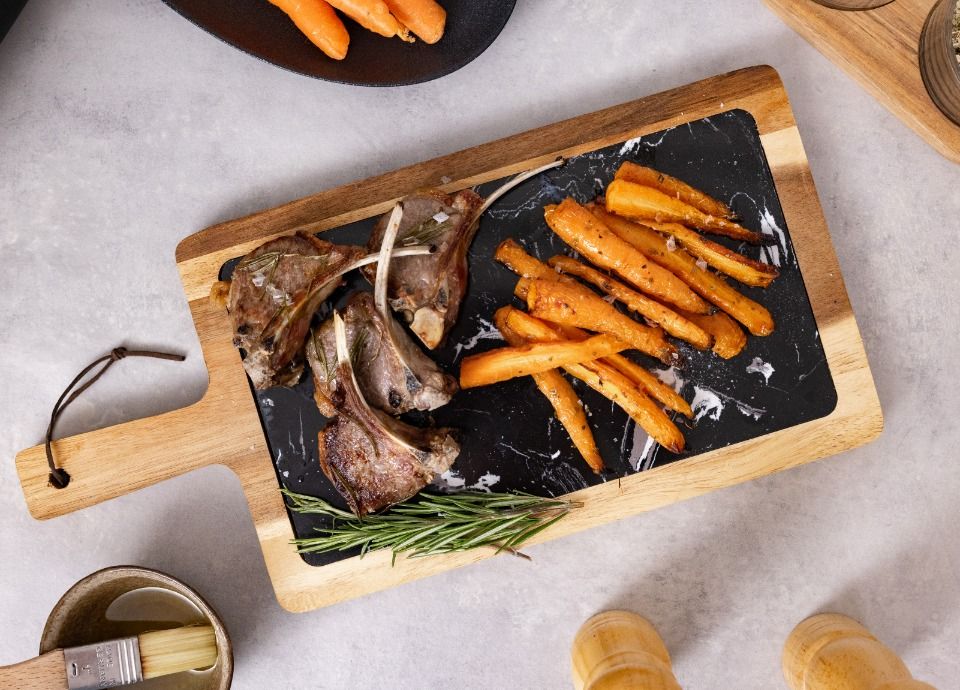 Grilled lamb chops served with roasted carrots on a wooden serving board, garnished with rosemary and presented on a light stone surface.