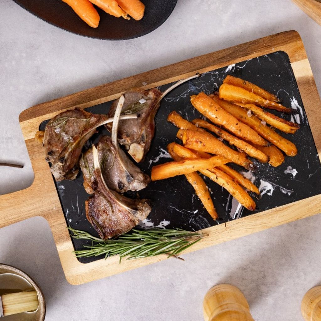 Grilled lamb chops served with roasted carrots on a wooden serving board, garnished with rosemary and presented on a light stone surface.