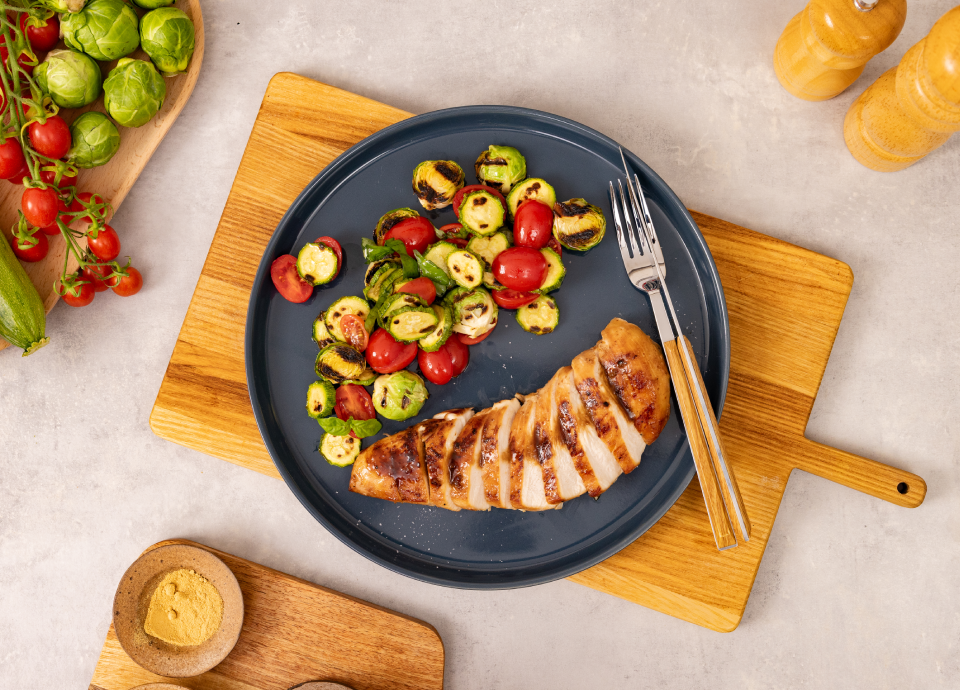 Petto di pollo grigliato a fette servito con zucchine, cavoletti di Bruxelles e pomodorini su un piatto scuro.
