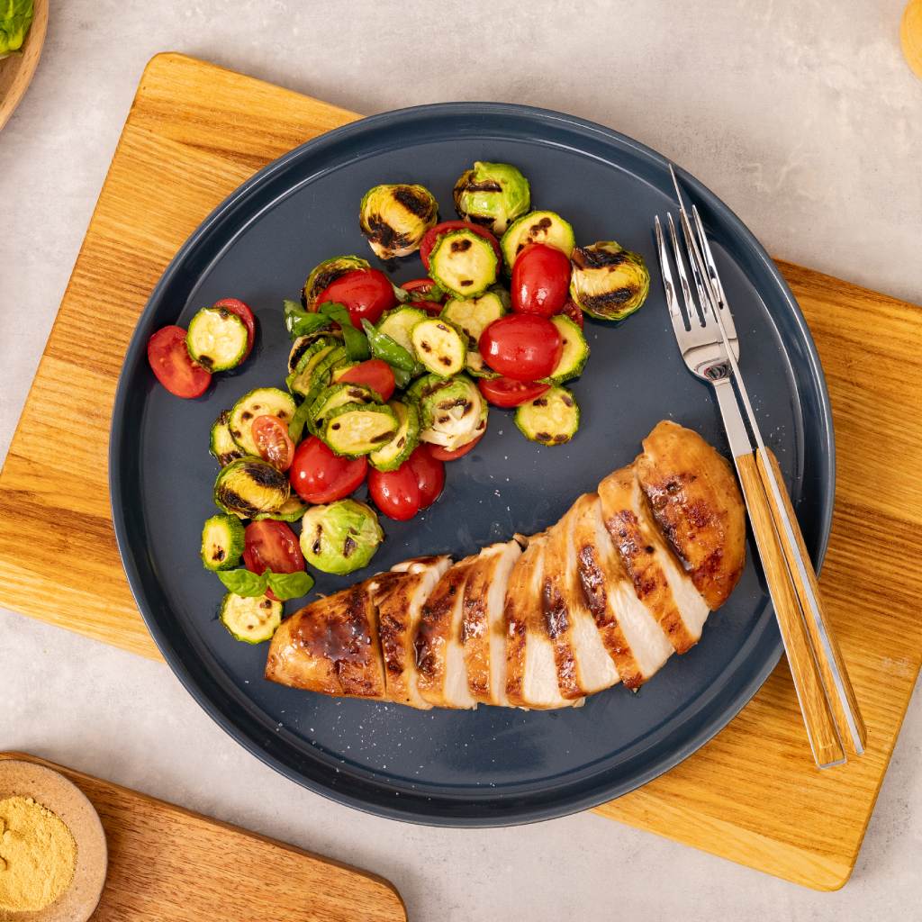 Petto di pollo grigliato a fette servito con zucchine, cavoletti di Bruxelles e pomodorini su un piatto scuro.