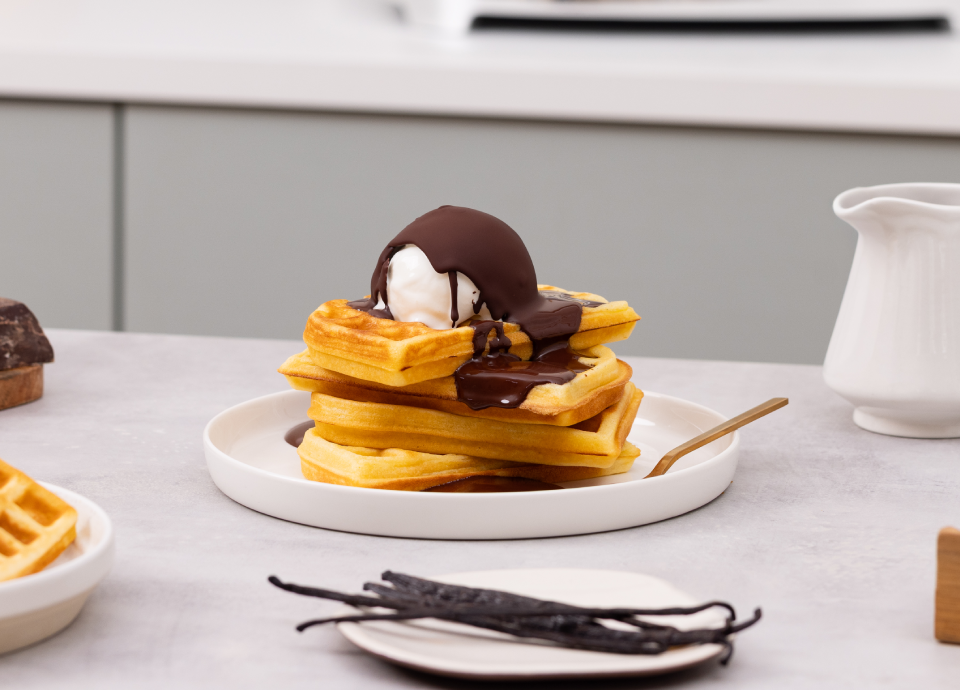 Pila di waffle dorati con gelato alla vaniglia e salsa al cioccolato fuso su un piatto bianco.