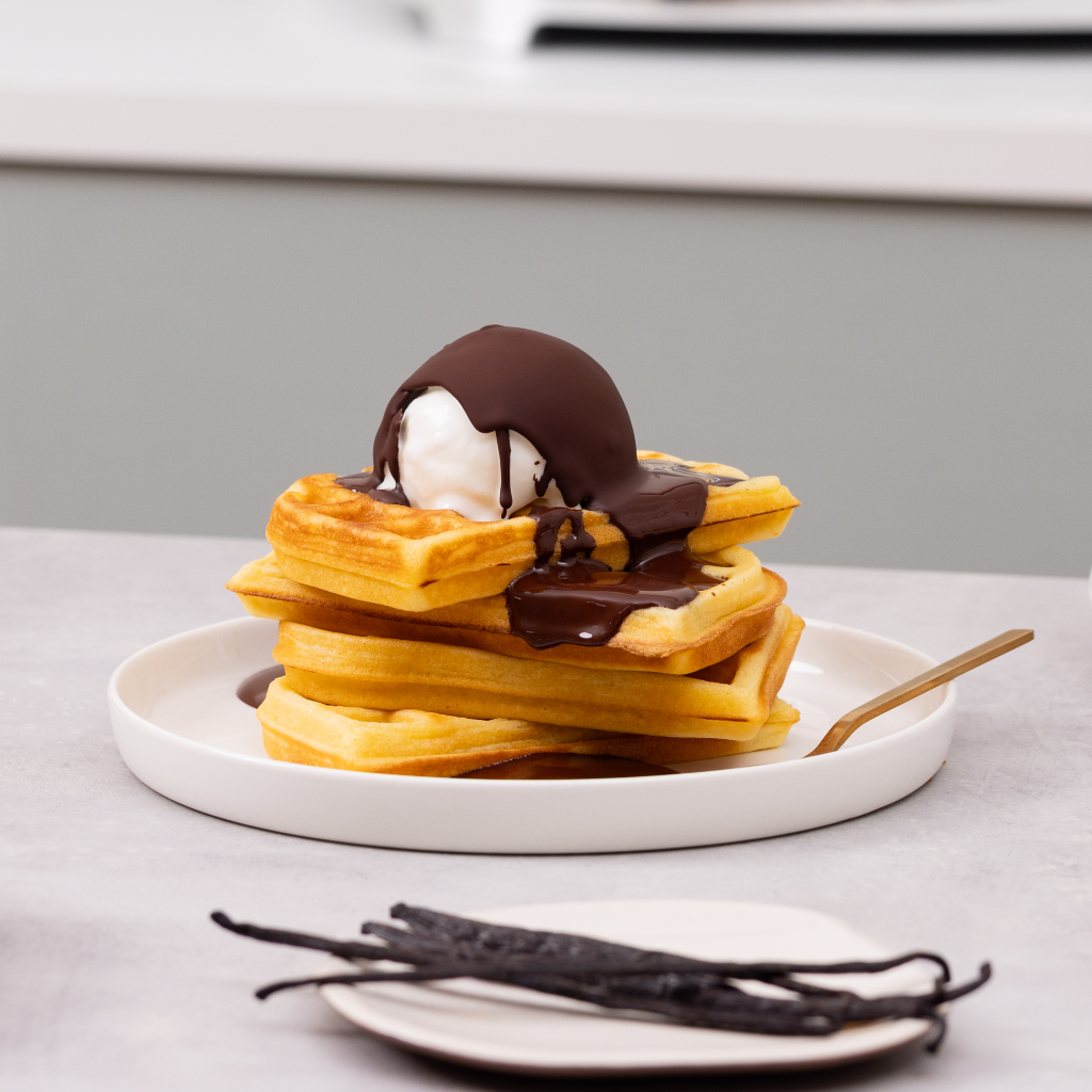 Pila di waffle dorati con gelato alla vaniglia e salsa al cioccolato fuso su un piatto bianco.