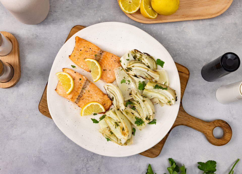 Filetti di salmone alla griglia serviti con finocchi arrosto e fette di limone su un piatto bianco.