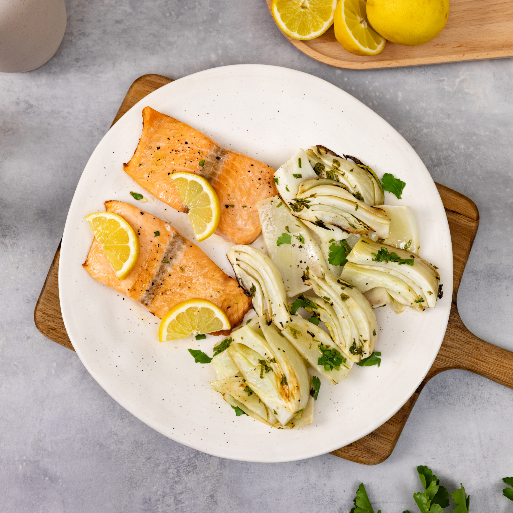 Filetti di salmone alla griglia serviti con finocchi arrosto e fette di limone su un piatto bianco.