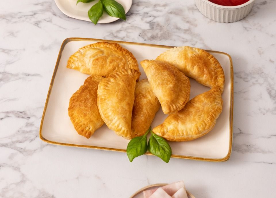 Golden puff pastry panzerotti served on a plate, with mozzarella, tomato sauce, fresh basil and ham on a marble countertop.