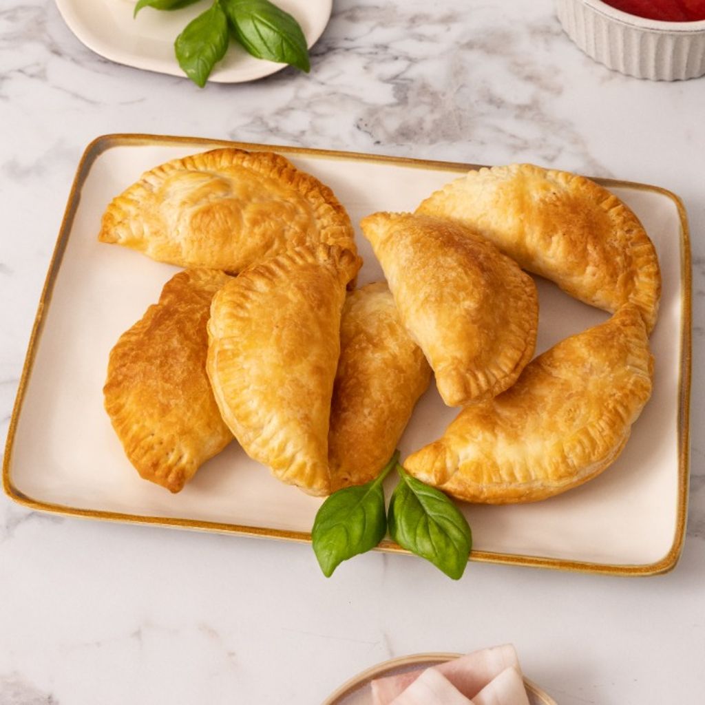 Golden puff pastry panzerotti served on a plate, with mozzarella, tomato sauce, fresh basil and ham on a marble countertop.