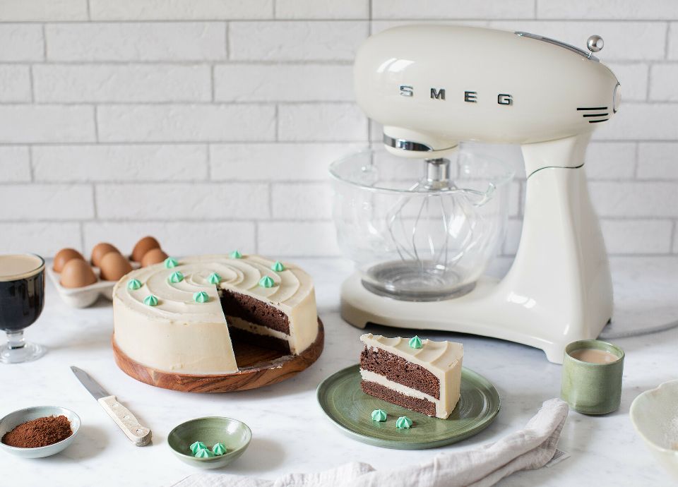 A two-layer chocolate cake with pale frosting and green piping, next to a cream Smeg stand mixer with a glass bowl.
