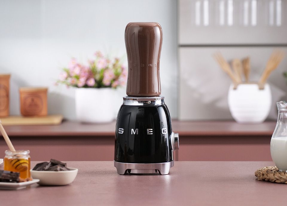 Black Hand blender on a kitchen counter, with jars, chocolate pieces, and flowers in the background.