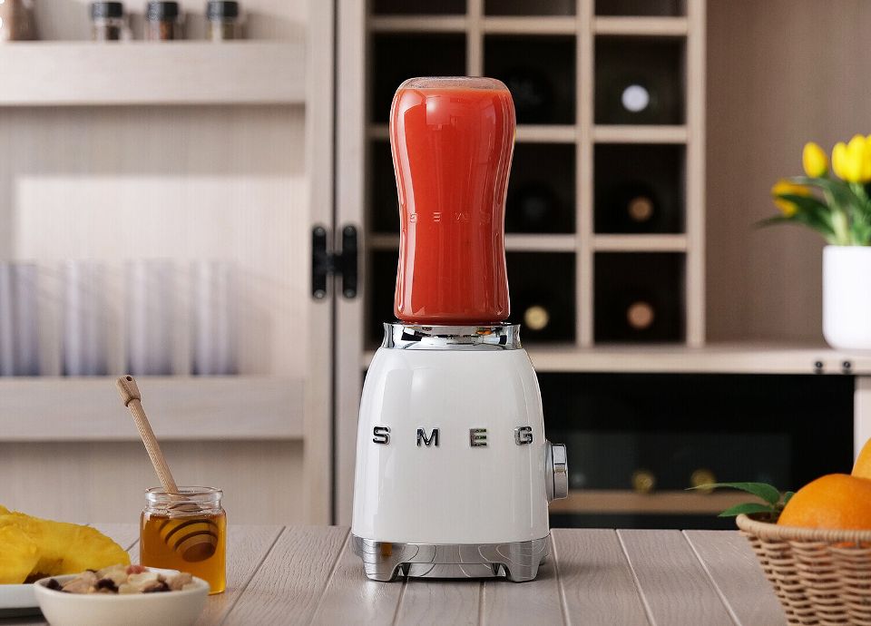 Frullatore a immersione bianco con smoothie rosso su un piano cucina, con barattolo di miele, arance e una pianta sullo sfondo