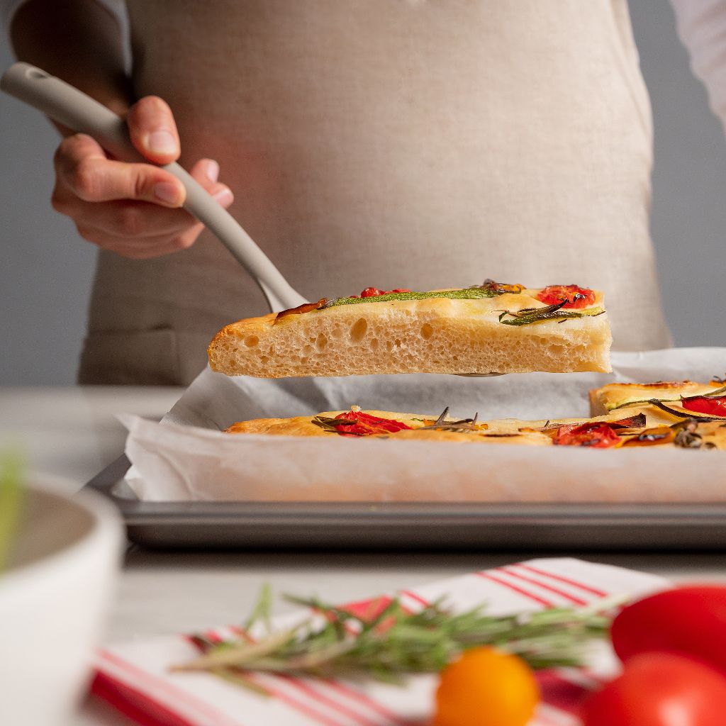 Fetta di focaccia con pomodorini ed erbe sollevata con una spatola sopra una teglia ricoperta di carta da forno.